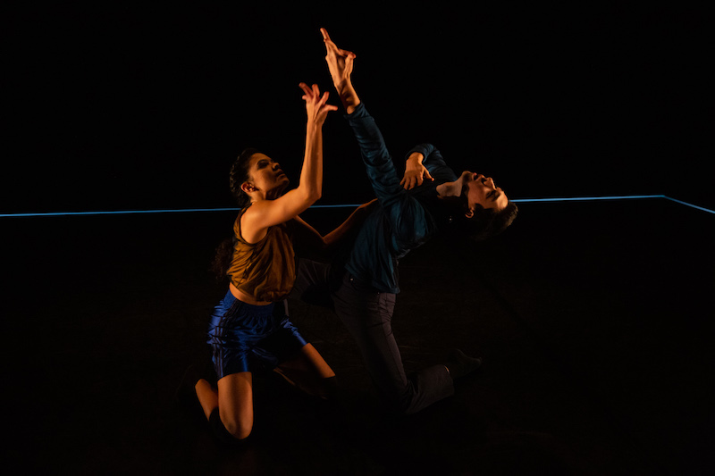 Two dancers, in semi darkness, kneel facing one another and reach each of their arms to the ceiling.